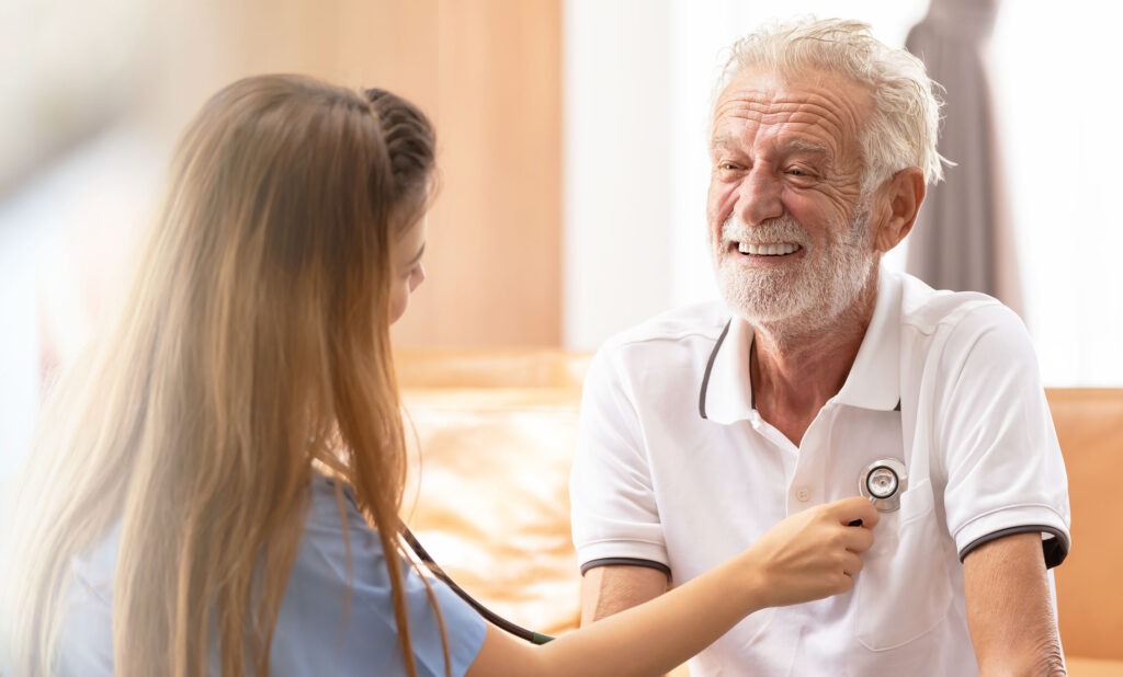 a healthcare professional, likely a nurse, attending to an elderly gentleman. The nurse is in the process of listening to the man's heart, using a stethoscope, which suggests a routine health check-up. The atmosphere is light-hearted and friendly; the older man is smiling broadly, showing his contentment and trust in the care he's receiving. The nurse, though her back is to the camera, seems to be focused on her patient, indicating a professional and caring interaction. The setting appears to be a comfortable room with soft lighting, adding to the sense of a personalized and attentive healthcare experience.